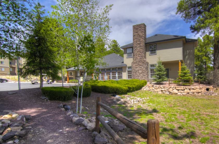 A tan two-story house with a stone chimney sits beside a small garden at Highland Village Apartments Flagstaff, surrounded by trees, bushes, rocks, and a wooden fence under a partly cloudy sky. A paved walkway and parked cars are visible in the background.