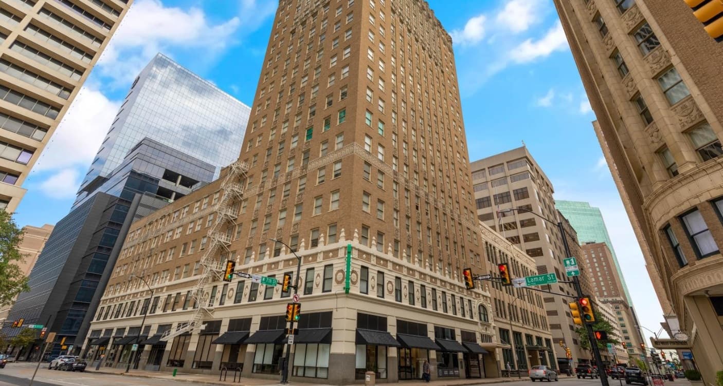 The tall, historic brown brick building is the Historic Electric Building Apartments Fort Worth, standing proudly at a busy city intersection amid modern skyscrapers under a blue sky with scattered clouds. Traffic lights and street signs frame the scene.