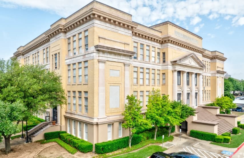 A large, historic, yellow-brick building with tall columns and many windows, reminiscent of the Homes of Parker Commons Fort Worth. Trees line the sidewalk and steps lead up to the entrance under a mostly clear sky with a few clouds.