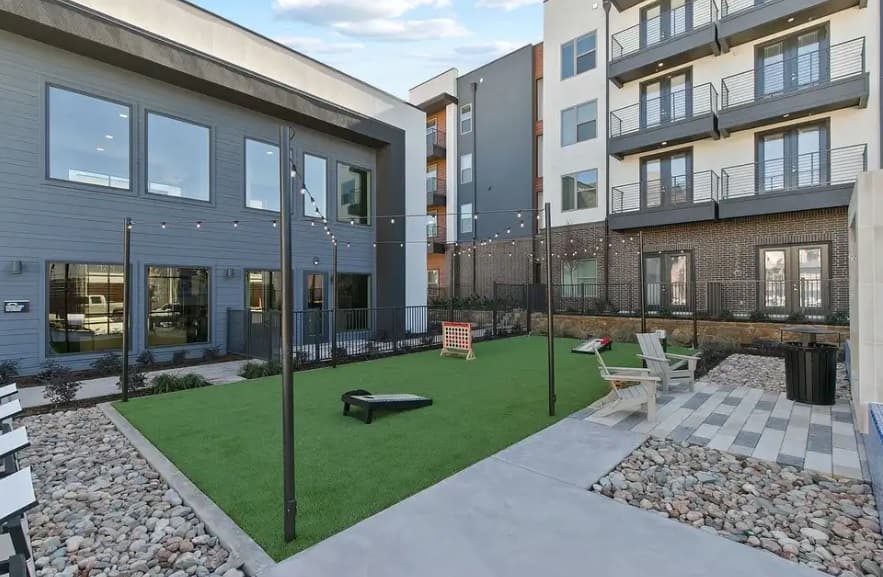 Apartment courtyard at Jameson at the Bluffs Fort Worth with artificial grass, string lights overhead, cornhole and Connect Four games, surrounded by modern buildings, outdoor seating areas, and rock landscaping.