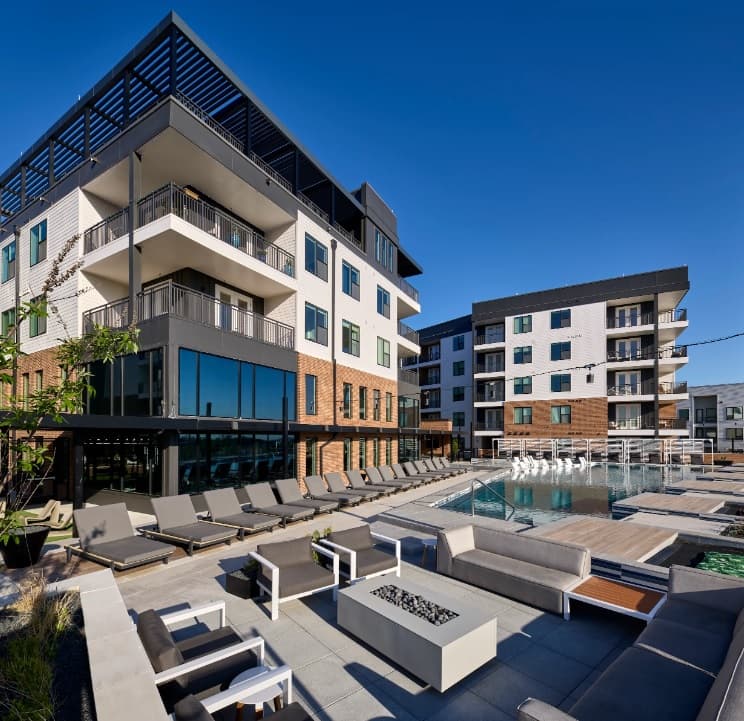 Modern apartment complex, Bexley at Left Bank Fort Worth, features multiple stories, balconies, and large windows. Enjoy the outdoor lounge with cushioned seating, fire pits, and a sparkling pool surrounded by lounge chairs under clear blue skies.