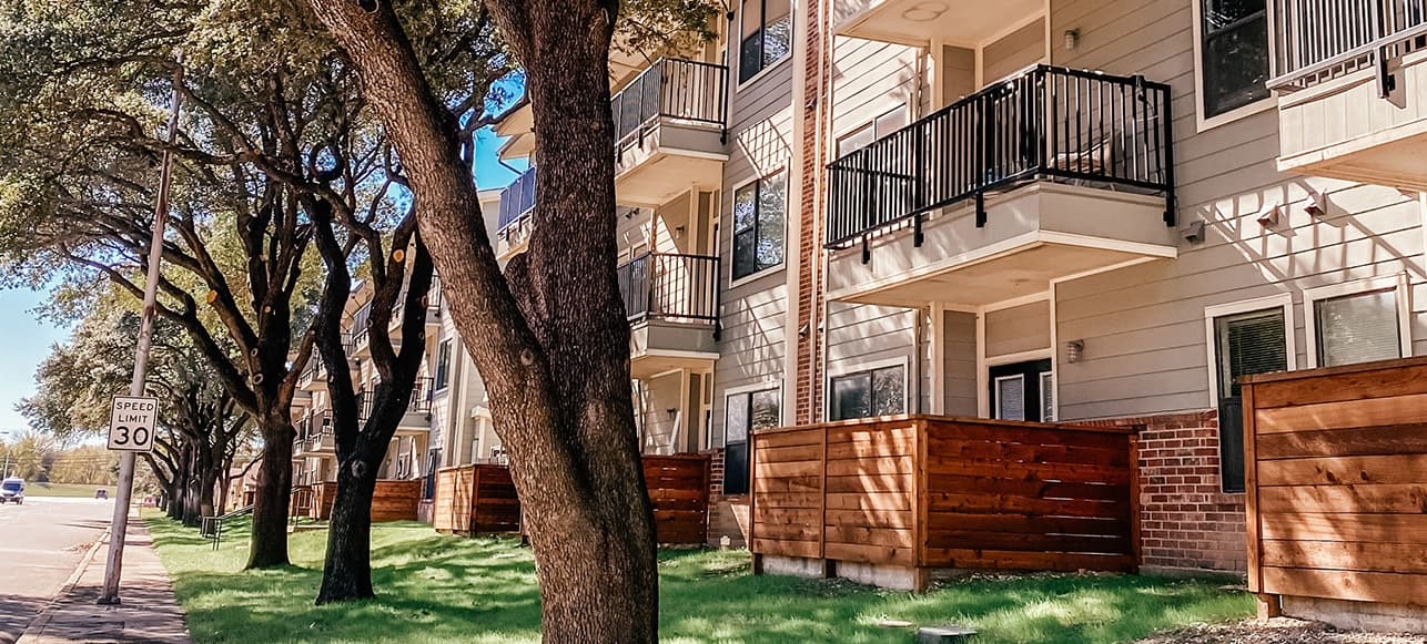 A row of modern apartment buildings, such as Liberty Lofts Fort Worth, with balconies and wooden fences shaded by tall trees, lines a sunny street with a 30 mph speed limit sign.