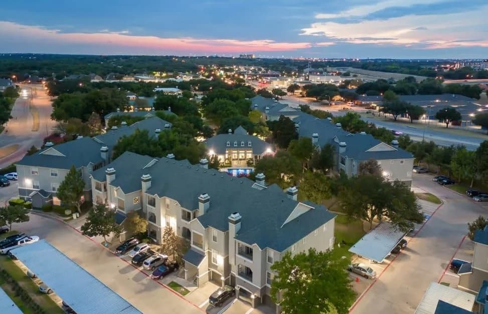 Aerial view of Marquis at Stonegate Fort Worth, a residential apartment complex with multiple gray-roofed buildings, surrounded by trees and parking areas, at dusk with city lights and a colorful sky in the background.