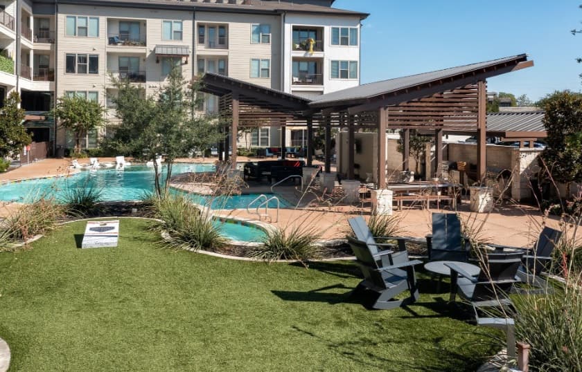 A modern apartment complex in Mercantile River District Fort Worth features a swimming pool, shaded lounge areas with chairs, a covered patio, and landscaped green spaces under a sunny sky.