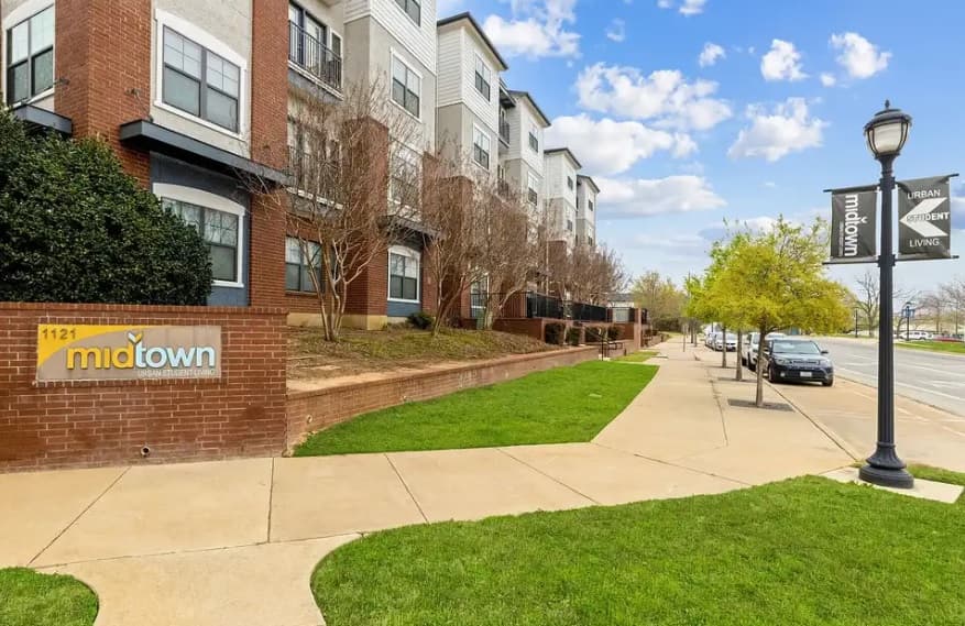 A sidewalk runs beside a modern apartment building with a brick sign reading 1121 Midtown, ideal for urban student living in Arlington. Trees and green grass line the street, with cars parked along the curb under a sunny, partly cloudy sky.