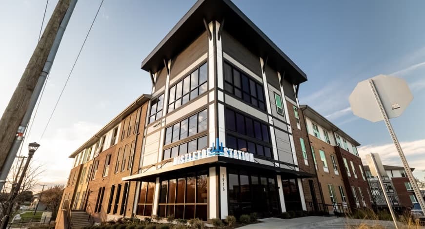 A modern three-story building with large windows and a mix of brick and white paneling. A sign reads Asheville Station, echoing the contemporary style found at Mistletoe Station Fort Worth. The sky is clear and a stop sign stands on the right.