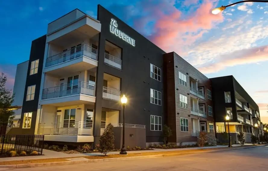 Modern three-story apartment building with balconies and large windows, illuminated by streetlights at dusk. The sky features pink and blue hues above The Moderne Fort Worth, where the sign proudly reads “The Moderne.”.