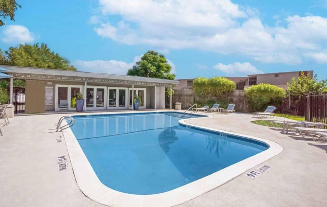 Outdoor swimming pool with clear blue water at Monticello Crossroads Fort Worth, surrounded by lounge chairs and trees. A small building with large windows is in the background under a partly cloudy sky. Depth markings line the pool’s edge.