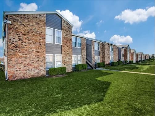 A row of two-story brick and gray-paneled Monarch Pass Apartments Fort Worth buildings stands with trimmed bushes and green grass under a blue sky with scattered clouds.