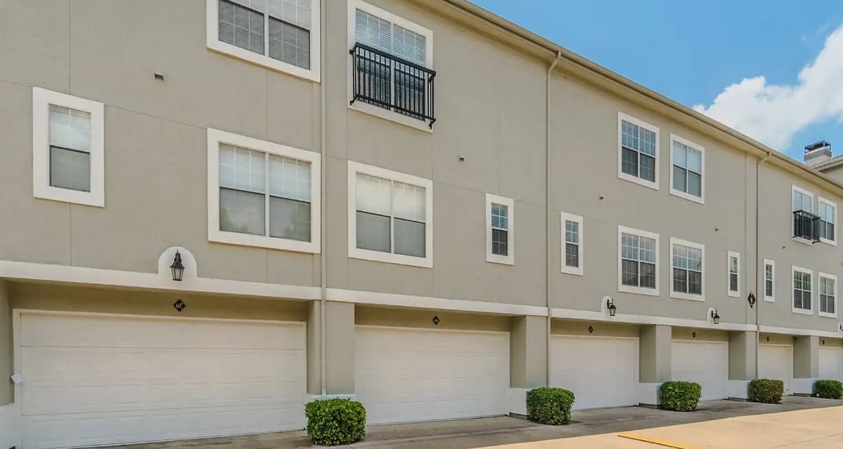 A row of modern beige townhouses at Monticello Oaks Townhomes Apartments Fort Worth, each with white trim, double garages, multiple rectangular windows, and small trimmed bushes in front, all under a clear blue sky.