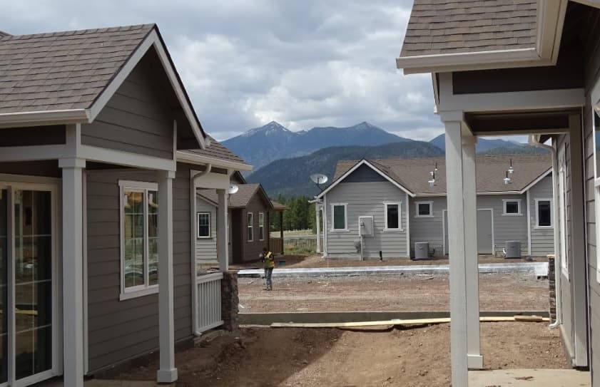 A row of gray houses with white trim is under construction at The Overlook at Buffalo Park Flagstaff, with a worker in a yellow vest in the distance. Snow-capped mountains and a cloudy sky form the scenic backdrop.