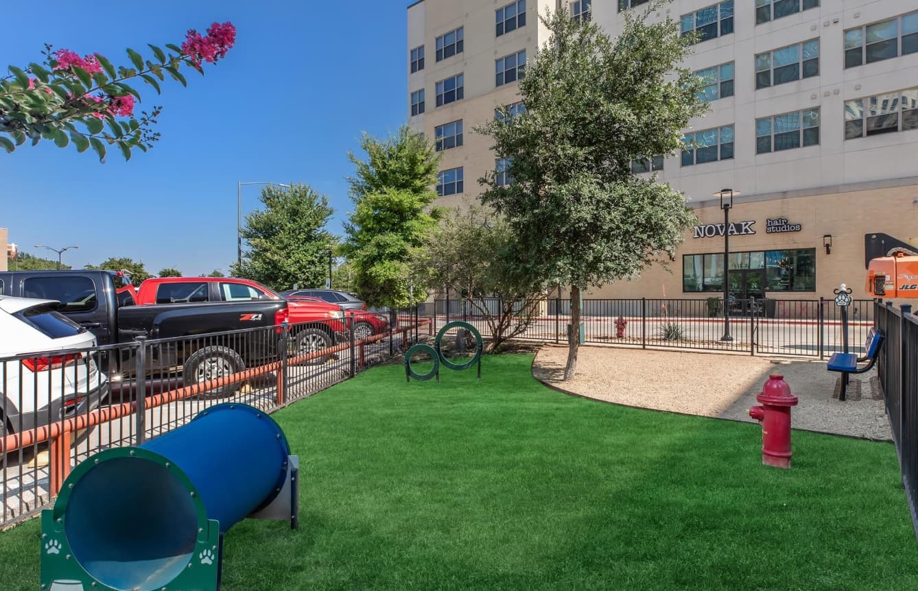 A small fenced dog park with green artificial grass, agility tunnels, hoops, a fire hydrant, and benches sits beside the parking lot near Parker House Fort Worth and a tall office building on a sunny day.