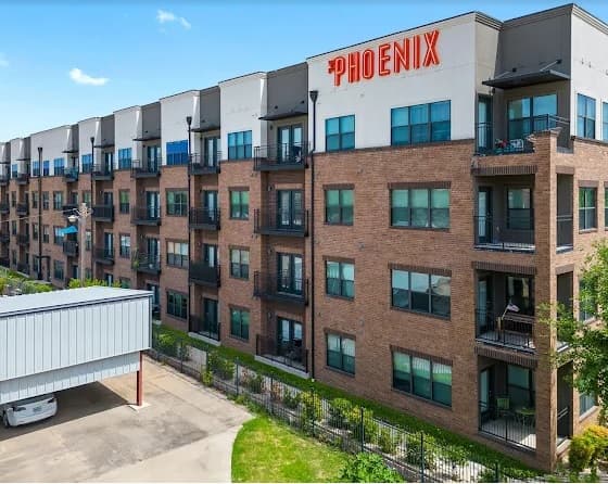 A modern four-story apartment building, The Phoenix Fort Worth features a brick and white exterior, balconies, and large windows. Enjoy covered parking and lush greenery along the fenced property line.