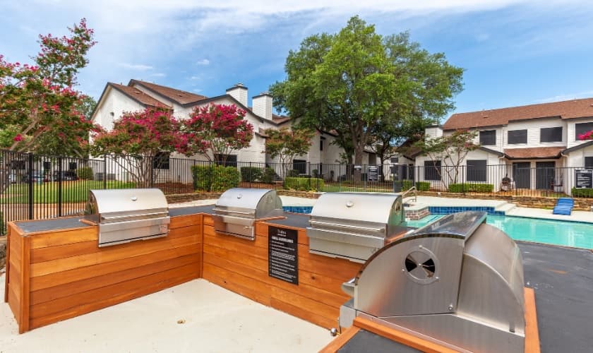 Four stainless steel grills are built into a wooden counter near a swimming pool at Prescott Park Apartment Homes Fort Worth, with townhouses, trees, and flowering bushes in the background under a partly cloudy sky.