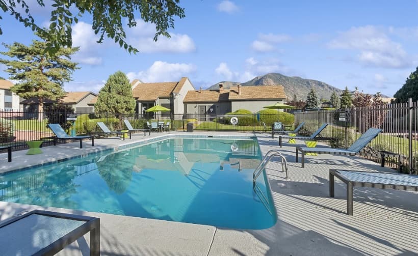 Outdoor swimming pool at ReNew Flagstaff East, surrounded by lounge chairs and umbrellas, with green bushes, trees, and residential buildings in the background. A mountain rises in the distance under a partly cloudy Flagstaff sky.
