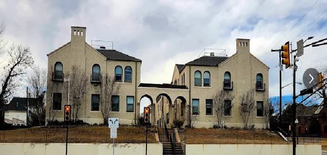 The Sasha at University Fort Worth is a beige building with two main sections connected by an archway, featuring arched windows and small balconies. Leafless trees and traffic signals are visible in the foreground under a cloudy sky.