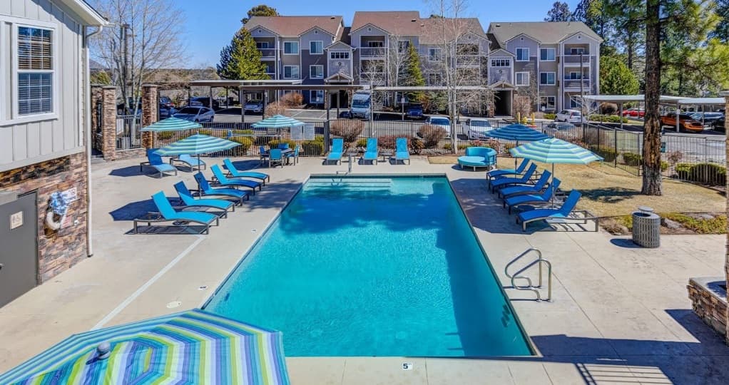 A rectangular outdoor swimming pool at Sterling Pointe Flagstaff is surrounded by blue lounge chairs and striped umbrellas, with apartment buildings, parked cars, and trees in the background on a sunny day.