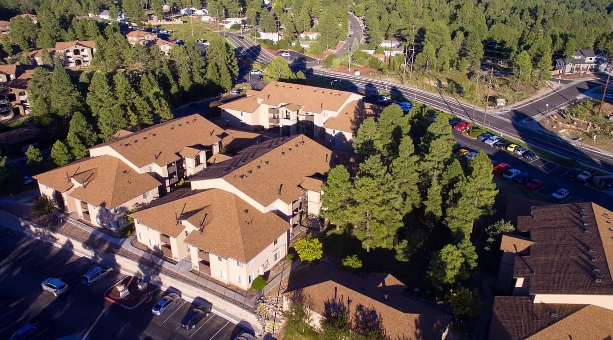 Aerial view of Table Rock Apartments Flagstaff, a residential complex with multiple tan buildings nestled among pine trees, next to several roads and parking lots in a suburban area.