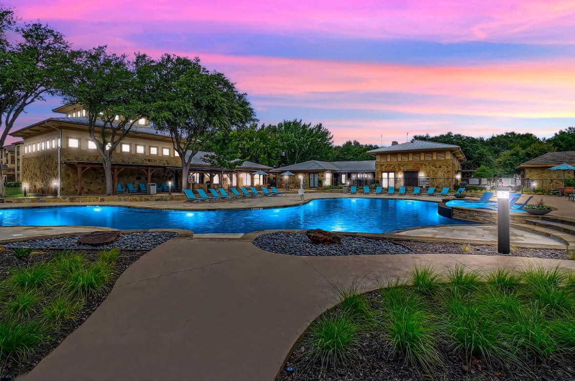 A resort-style outdoor swimming pool at The Canyons Fort Worth, surrounded by lounge chairs, lush greenery, and stone buildings, with trees and a vibrant pink and purple sunset sky in the background.