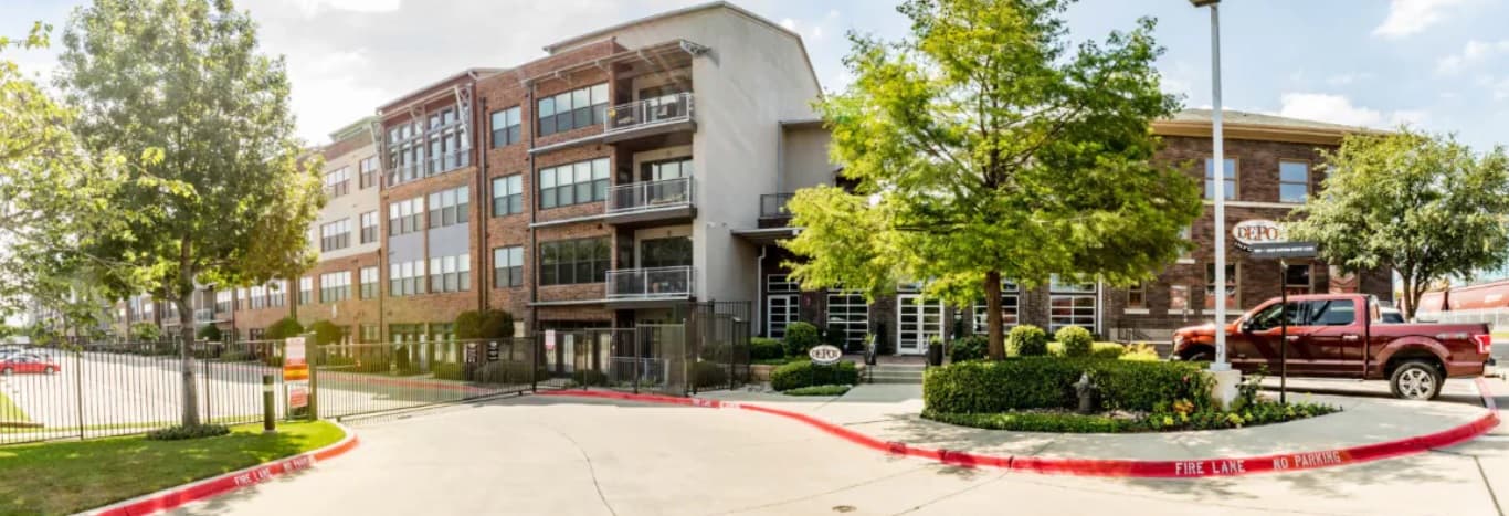 A modern, multi-story apartment building with brick and gray siding sits beside a tree-lined street near The Depot Fort Worth. A red pickup truck is parked by the entrance, and there’s a gated driveway and sidewalk in front.