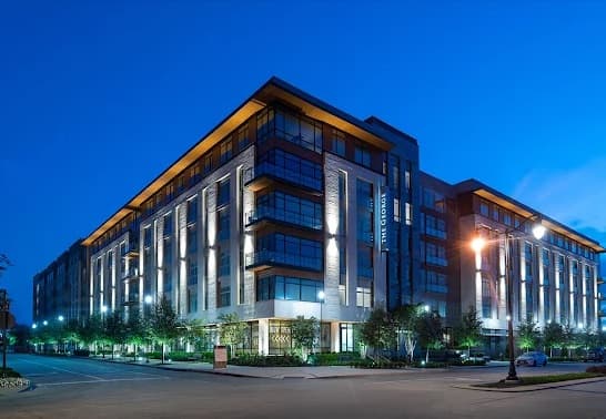 Modern, multi-story apartment building with large windows and illuminated exterior lights at dusk, The George Fort Worth stands surrounded by trees and street lamps on a quiet, empty street corner.
