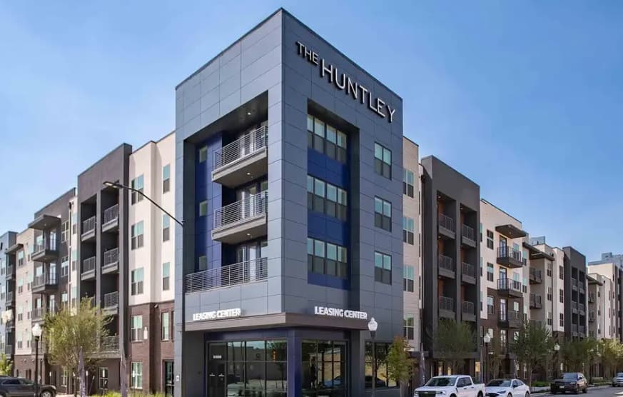 Modern apartment building The Huntley Fort Worth features a leasing center at ground level, spacious balconies, large windows, and several parked cars in front, all under a clear blue sky.
