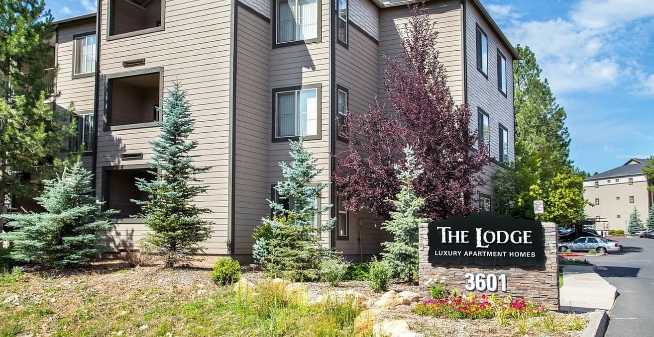 A brown, three-story apartment building with balconies and large windows sits behind landscaped greenery and a black sign reading The Lodge Luxury Apartment Homes Flagstaff 3601 on a sunny day.