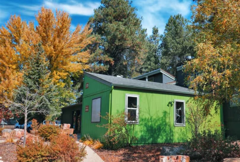 A small green house at The Place at Forest Ridge Flagstaff sits among colorful autumn trees and shrubs under a bright blue sky with scattered clouds. A pathway leads past rocks and plants in the front yard.