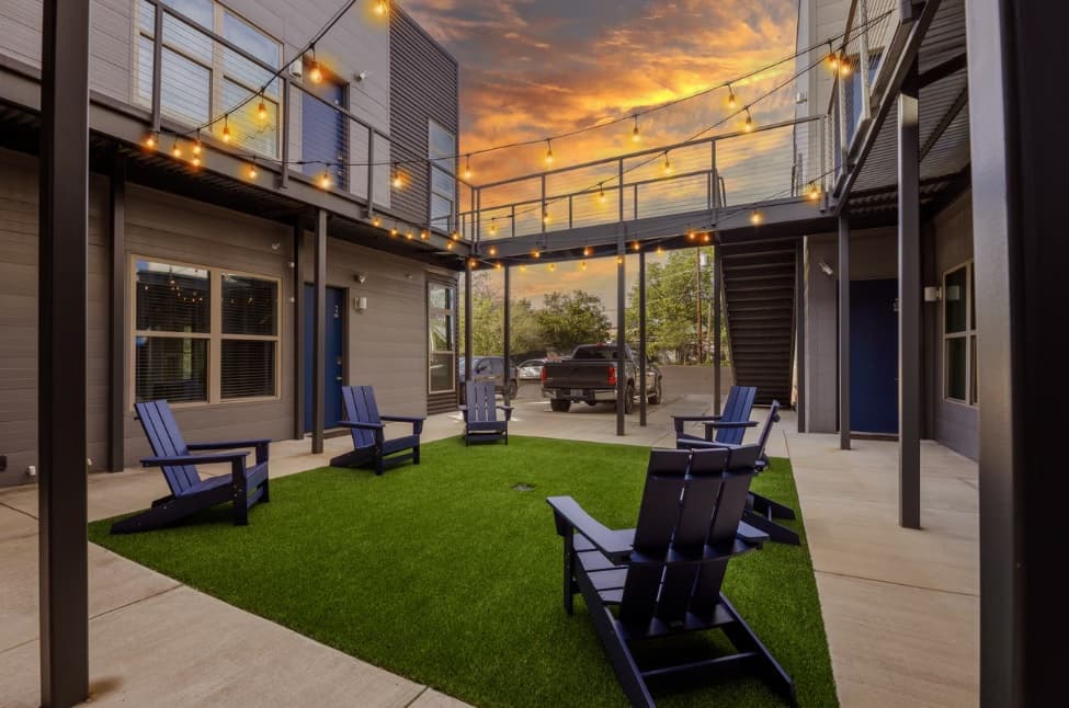 A modern outdoor courtyard at The Sasha at Jennings Fort Worth features blue Adirondack chairs on artificial grass, string lights above, and a two-story building with metal railings. A pickup truck is parked outside beneath the orange sunset sky.