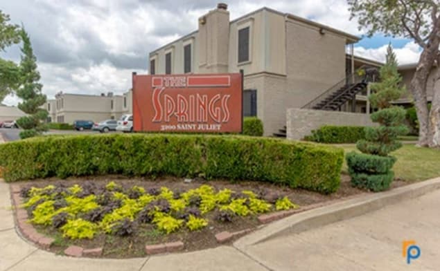 A red sign reading “The Springs, 2500 Saint Juliet” stands in front of a landscaped area with shrubs and flowers. The beige two-story apartment buildings of The Springs Fort Worth and a staircase are visible under a cloudy sky.