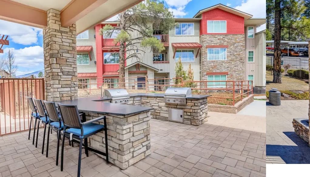 Outdoor patio area at Trailside Apartments Flagstaff features stone grill stations, countertop seating with blue chairs, and a view of the modern three-story building surrounded by trees and landscaped grounds.