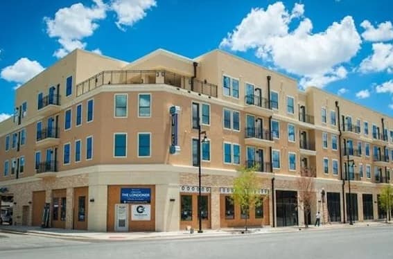 A modern, beige three-story apartment building near University House TCU Fort Worth, with many windows and balconies, shops on the ground floor, and a bright blue sky with scattered clouds in the background.