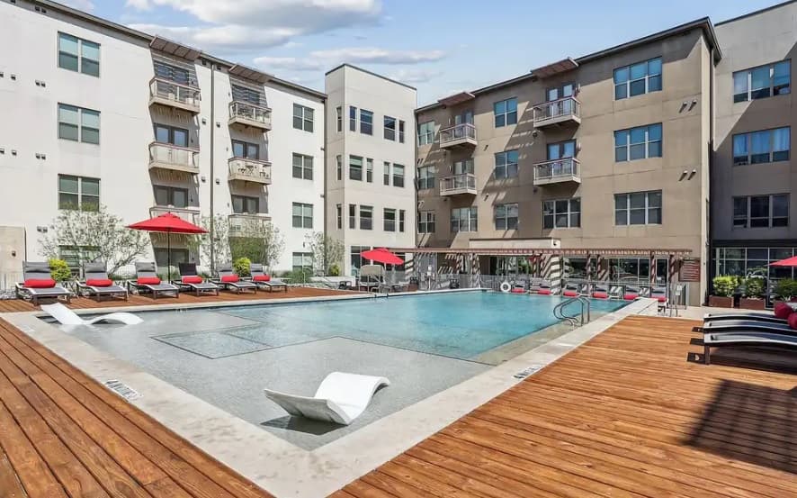 Modern courtyard at Parkside So7 Urban Apartments Fort Worth, featuring a rectangular outdoor pool, wooden deck with lounge chairs, red umbrellas, and multi-story buildings surrounding the pool under a partly cloudy sky.