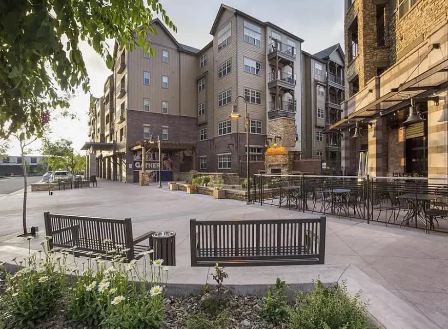 A modern apartment building at Village at Aspen Place Flagstaff features balconies overlooking a wide patio area with benches, outdoor seating, planters with flowers, and a lit outdoor fireplace, creating a welcoming urban atmosphere.