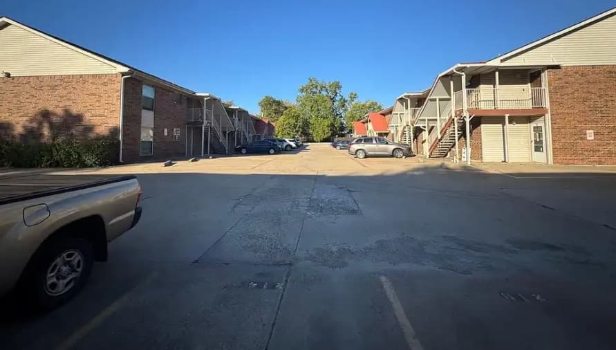 The Village East Apartment Fort Worth features a sunny parking lot flanked by two-story brick buildings, a few parked cars, and trees in the background under a clear blue sky.