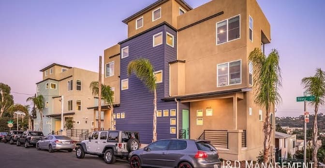 Modern three-story luxury townhomes with tan and blue exteriors line Riley Street, with parked cars and palm trees under a clear evening sky.