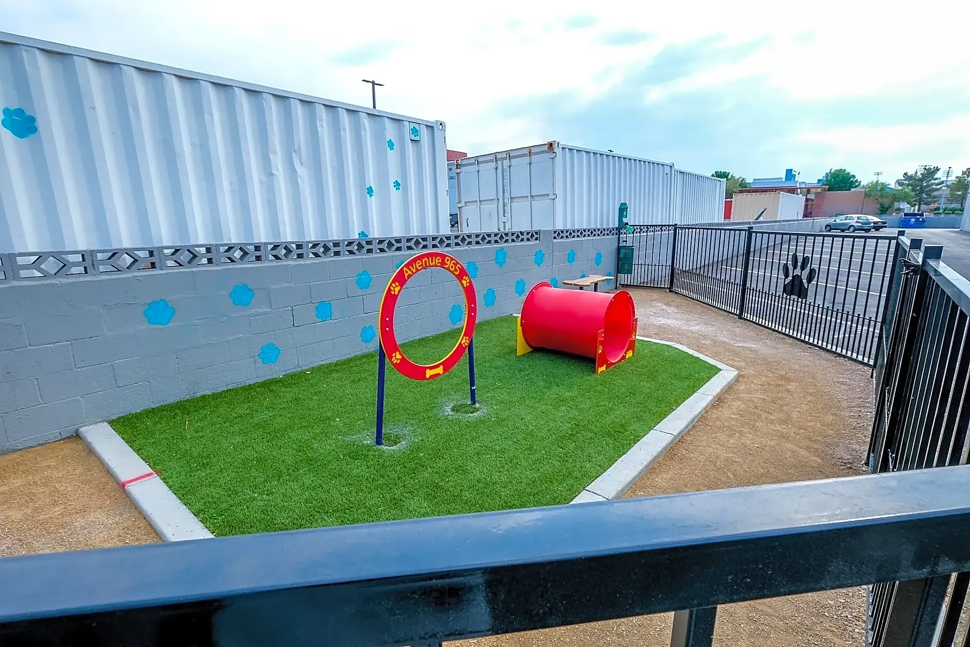 A small outdoor dog play area with artificial grass, a red agility hoop labeled K-9 Agility Spot, a red tunnel, a gray block wall with blue paw prints, and metal shipping containers in the background.
