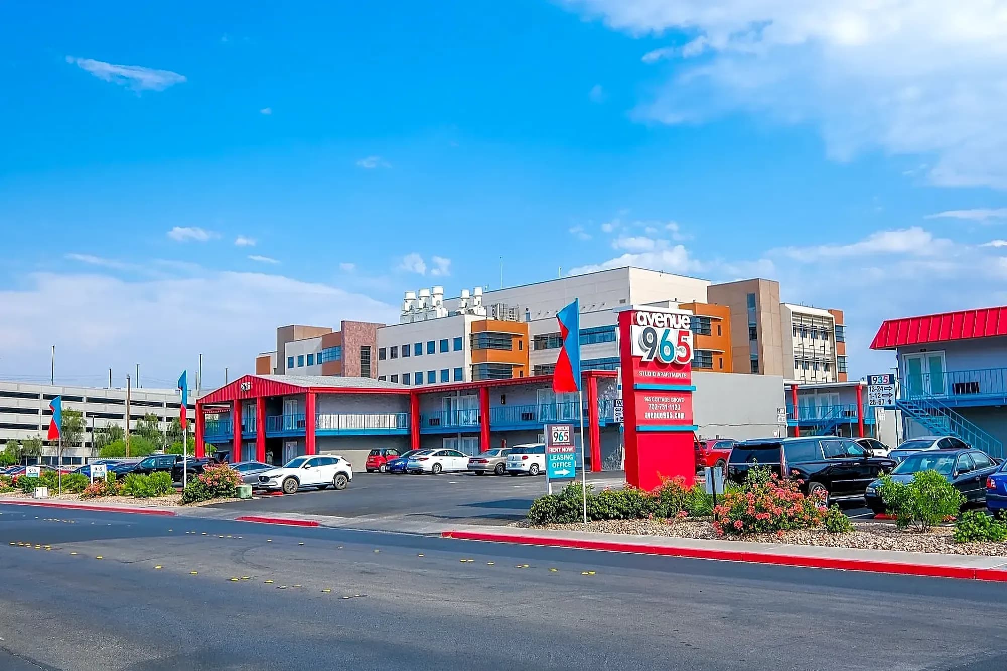 A mid-century style motel with red accents and a sign reading “OYO Hotel 965,” surrounded by parked cars, landscaped bushes, and a larger building and blue sky in the background.