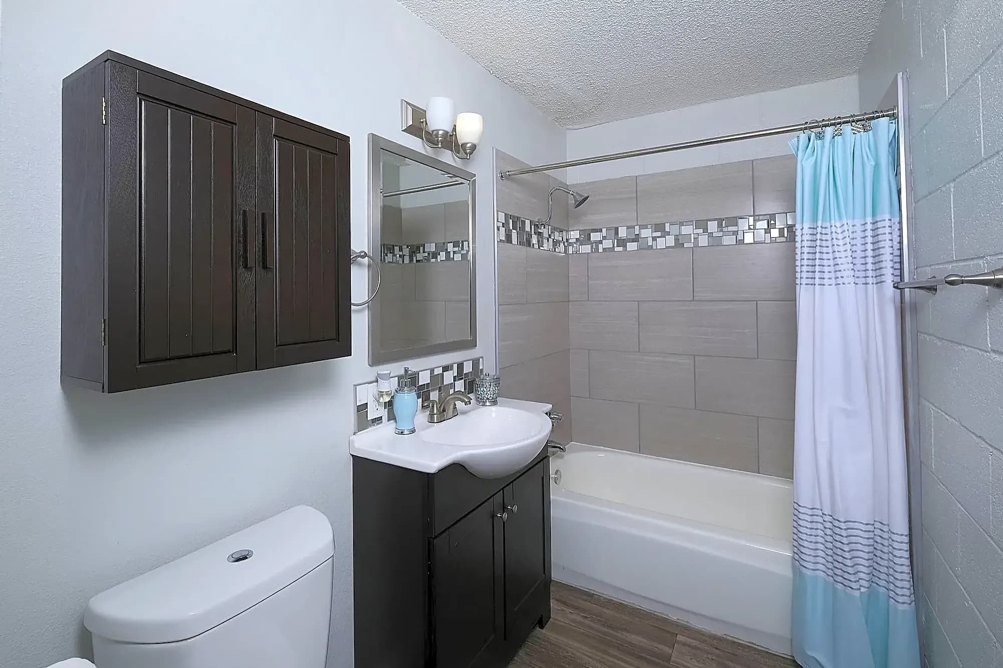 Modern bathroom with a dark wood vanity and cabinet, white sink and toilet, large rectangular wall tiles, a bathtub with a blue and white shower curtain, wood-look flooring, and contemporary lighting above the mirror.