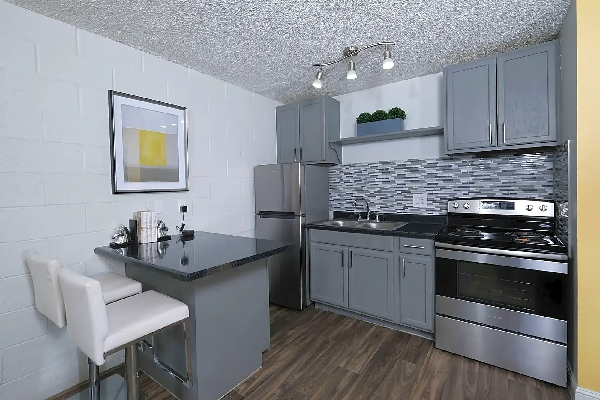 Modern kitchen with gray cabinets, stainless steel appliances, a tile backsplash, and a dark countertop. Two white cushioned chairs are at a small dining bar. A framed abstract art piece hangs on the wall.