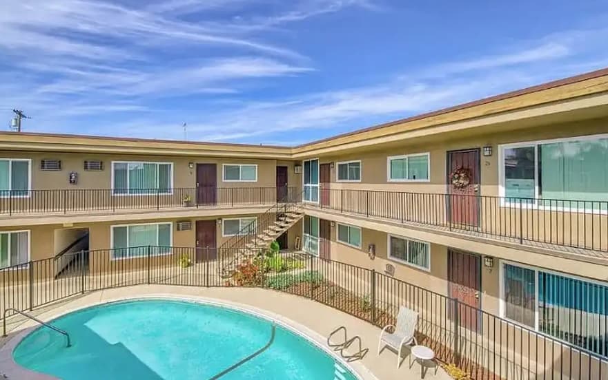 The Courtyard on 68th is a two-story apartment complex with beige walls and brown doors surrounding a small outdoor pool. Patio chairs line the pool area, and an exterior staircase leads to the upper rental level beneath clear, wispy clouds.