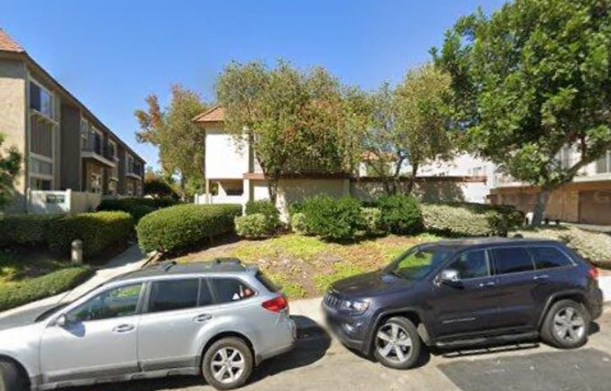 Two cars are parked on a residential street in front of a landscaped area with bushes and trees at 7798 Starling Dr. Behind them are two-story beige buildings with red-tiled roofs under a clear blue sky.