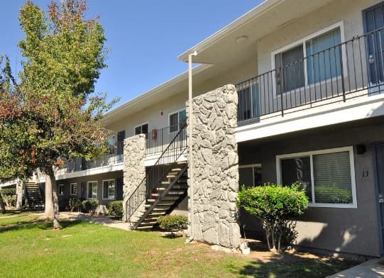 Vista Capri Apartments in Kearny Mesa is a two-story building featuring exterior stairs, stone-textured columns, and balconies. The property is surrounded by grass, bushes, and trees under a clear blue sky.
