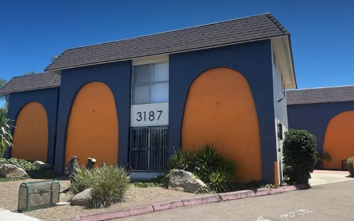 A two-story building with a dark blue exterior, large orange arch designs on its walls, and the number 3187 displayed beside a gated entry, surrounded by dry landscaping and a clear blue sky.