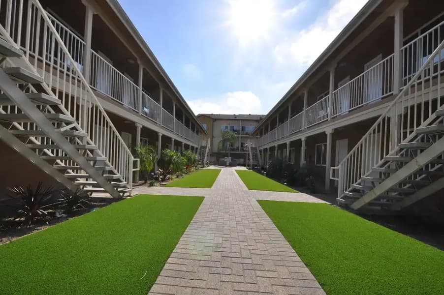 A courtyard at Astro Vista Apartments lies between two parallel, two-story buildings with staircases, railings, and artificial green grass on each side of a brick walkway, photographed on a sunny day.