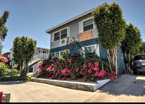 A two-story blue house at Aztec Lofts Apartments features a Now Leasing! sign, vibrant pink flowers, and tall trees. A driveway leads to the home with a parked car on the right under a clear, sunny sky.