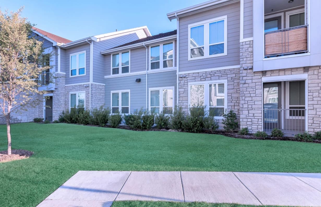 A modern Campus Apartments building with large windows, light gray siding, and stone accents is surrounded by neatly trimmed bushes, a green lawn, and a sidewalk in front.