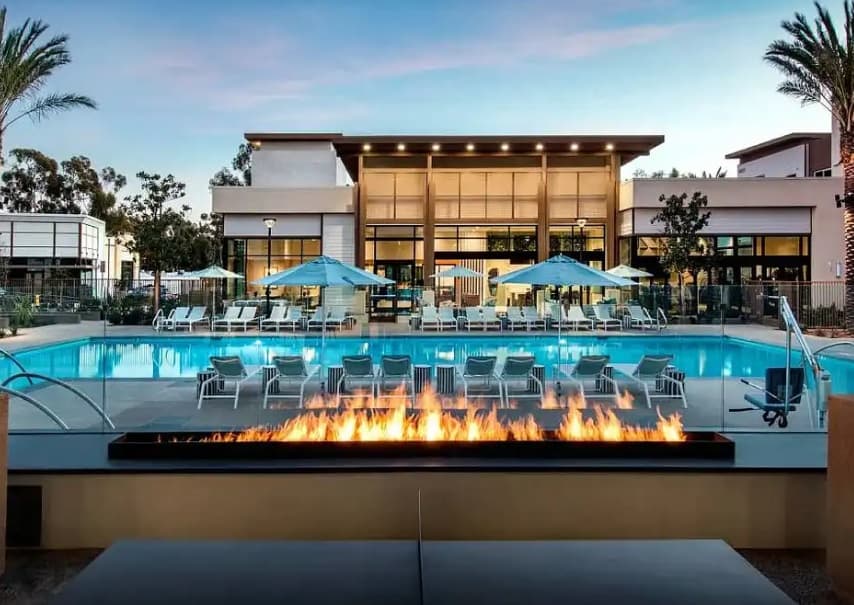 Modern outdoor pool area at 9870 Carroll Canyon Rd with lounge chairs and blue umbrellas, a rectangular fire pit in the foreground, and a contemporary building with large windows in the background at sunset.