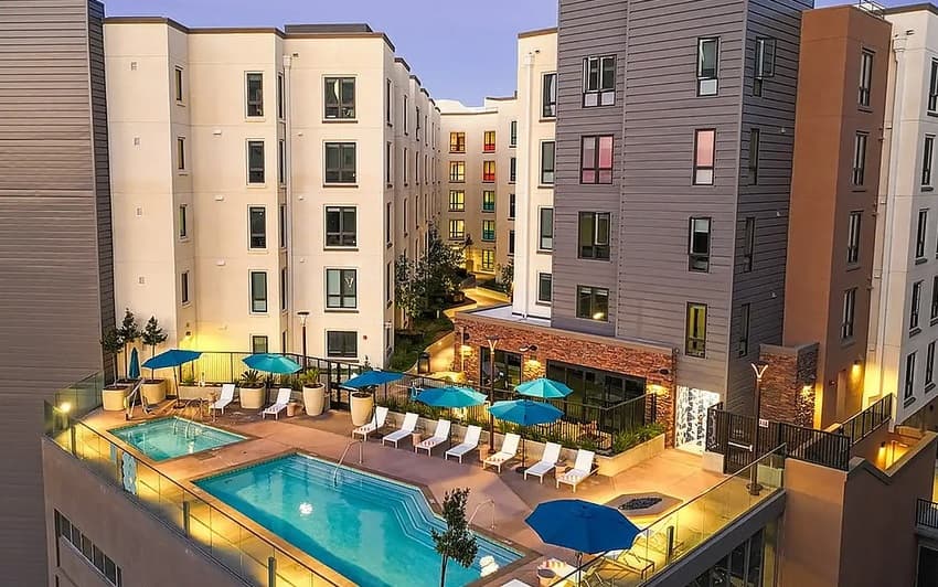 College View Apartments is a modern complex featuring a rooftop swimming pool, lounge chairs, and blue umbrellas. The several-story buildings have large windows and glowing outdoor lighting, creating an inviting atmosphere at dusk.
