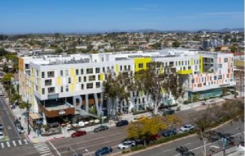 A modern multi-story apartment complex with white and yellow accents is situated at a street intersection with cars parked along the roads; the cityscape and blue sky are visible in the background.